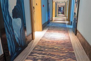 Een hotelgang met een kamerbreed tapijt, deuren en kunstwerken in lijsten aan de wanden.