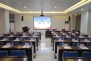 Un salón de conferencias con filas de mesas, sillas y una pantalla de proyector al frente.