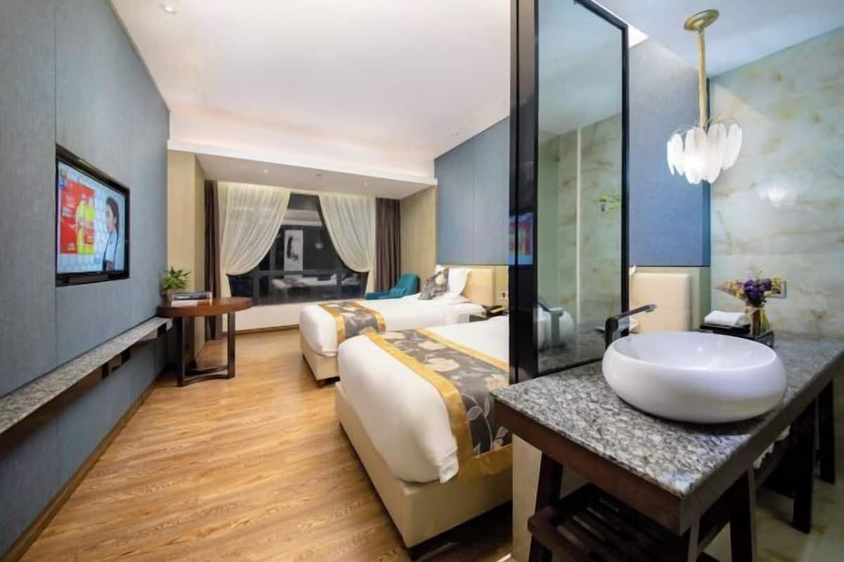 A hotel room with a large bed, a separate sofa, a small table, a large mirror, and a bathroom with a sink and shower.