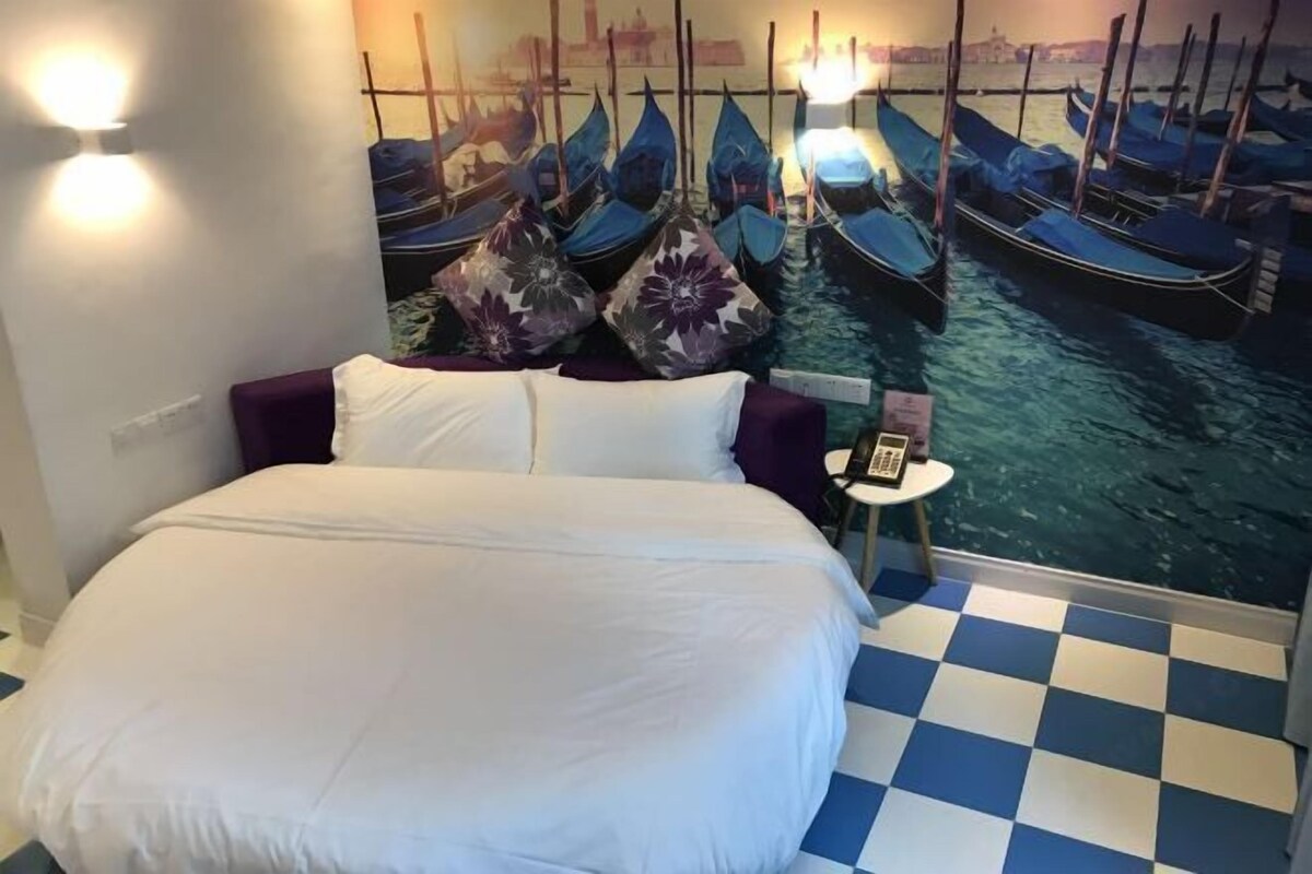 Ein Hotelzimmer mit einem großen Bett, einem Schachbrettfußboden, einem Wandbild mit Booten und einem kleinen Tisch mit Lampe.