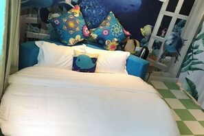 Ein Bett mit weißer Bettwäsche, einem blauen Kopfkissen mit Fischmuster und bunten Kissen mit Blumenmotiven.