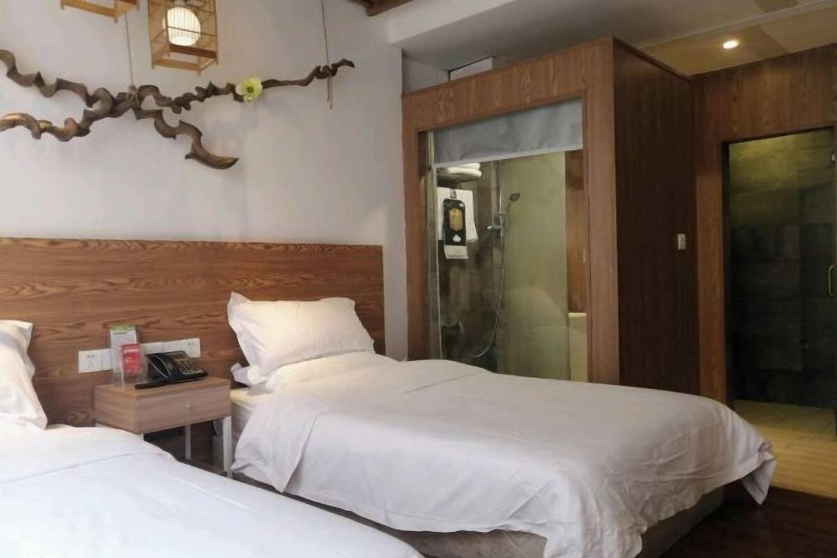 Habitación de hotel con dos camas, cabecero de madera, mesita de noche con teléfono y zona de ducha.