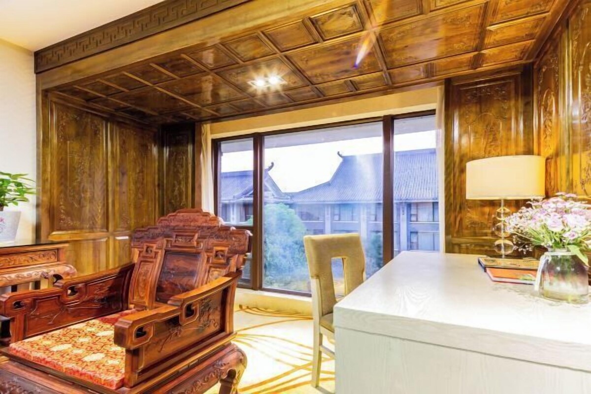 A room with a wooden ceiling, a large window with a view of buildings, a wooden chair, a white table, and a lamp.
