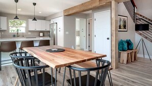 Une cuisine moderne avec des meubles blancs, une table à manger en bois et des chaises noires. On y trouve un escalier avec une rampe en bois et un tableau encadré au mur.