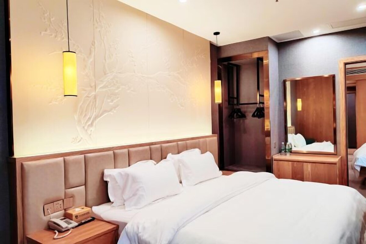 A hotel room with a large bed, a headboard with a telephone and a box, a bedside table with a lamp, and a mirror.