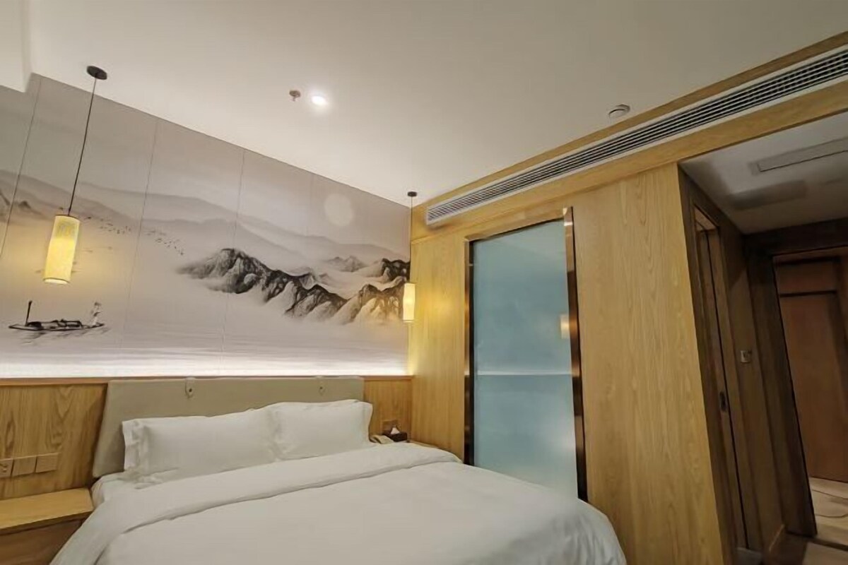 A hotel room with a bed, a wooden headboard, a wall-mounted artwork of mountains, and a hanging light fixture.