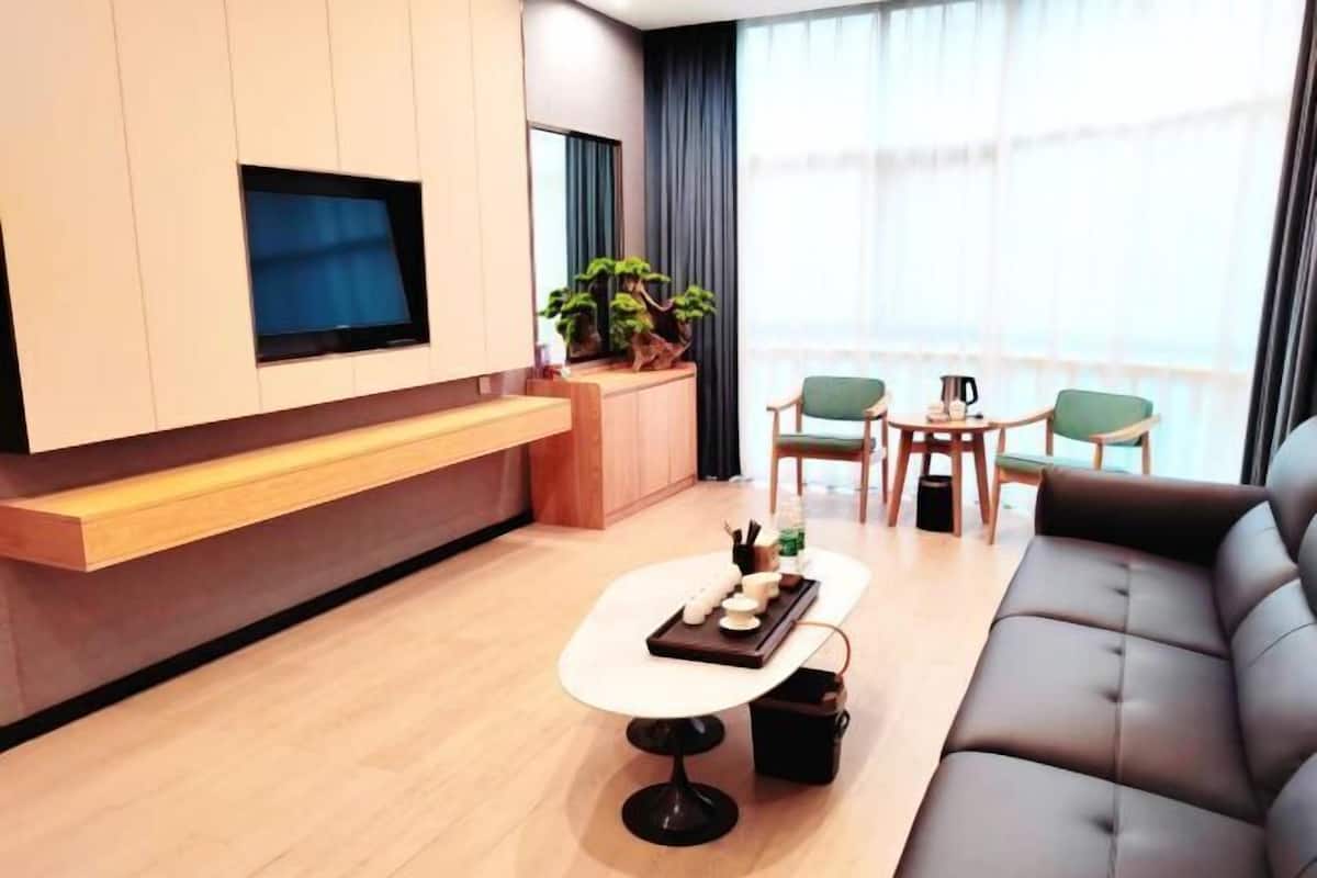 A modern living room with a sofa, a coffee table, and a TV mounted on the wall. There are two chairs and a small table with a plant and a coffee pot.