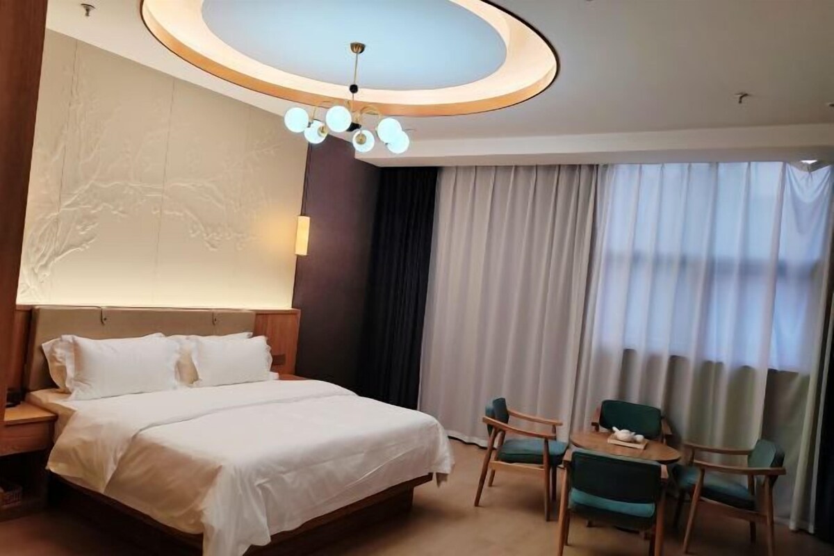 A hotel room with a large bed, a desk with a chair, a chandelier, and a view of the outside through large windows.
