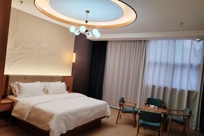 Une chambre d’hôtel avec un grand lit, un bureau avec une chaise, un lustre et une vue sur l’extérieur grâce à de grandes fenêtres.
