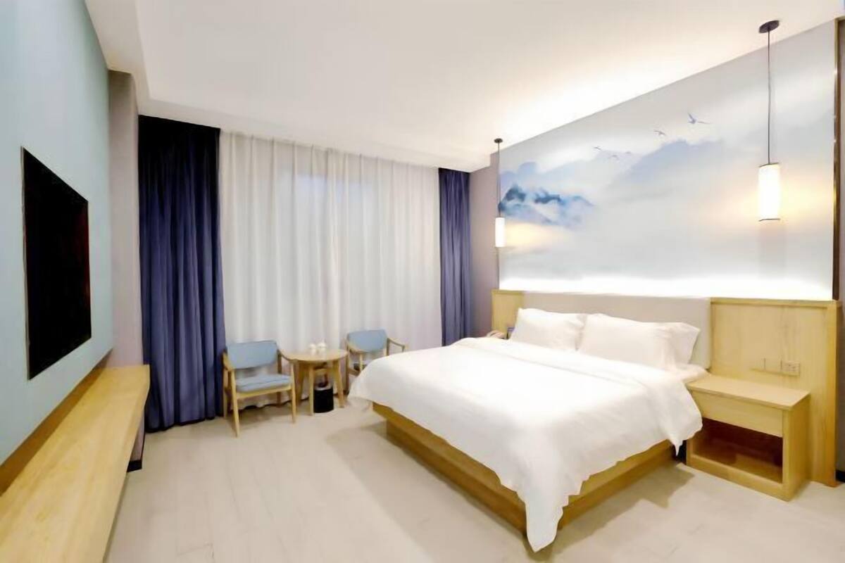 A hotel room with a large bed, a TV, a desk with a chair, and a wall with a cloud mural.