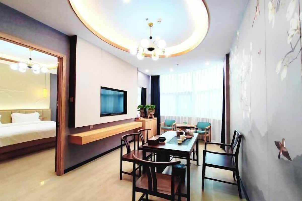 A modern hotel room with a bed, a TV, a dining area with chairs, a table, and a ceiling light fixture.