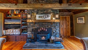 Un salon chaleureux avec une cheminée en pierre, des poutres apparentes et une bibliothèque remplie de livres et d’objets décoratifs.