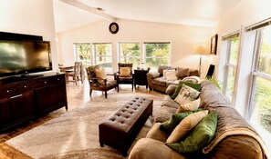 A spacious living room with a large flat-screen TV, a dark wooden entertainment center, a brown leather sofa, a matching ottoman, and a patterned armchair.