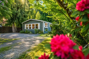A small house with a gravel driveway, surrounded by lush greenery and flowering plants.