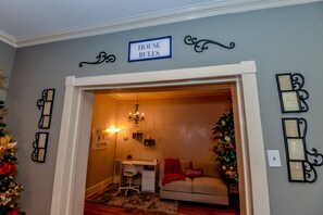 Quarto com escrivaninha, um pequeno sofá, um candelabro e um aviso com as regras da casa na parede.