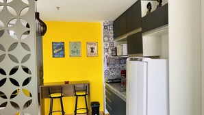 A compact kitchen with a yellow accent wall, a refrigerator, a microwave, and a small dining area with two stools. - Studio (RJ)