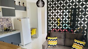 A modern living room with a brown sofa and two patterned cushions, a kitchen area with a refrigerator, a yellow chair, and a decorative wall with a geometric pattern. - Studio (RJ)