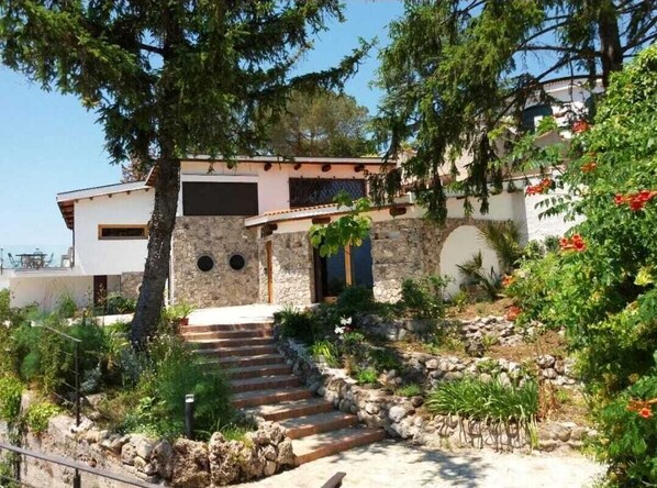 A stone house with a stone staircase leading to a wooden door, surrounded by greenery and a clear sky.