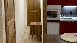 Un passadís estret amb portes de fusta i una petita banca de fusta. Se veu una cuina al fons amb armaris vermells i electrodomèstics blancs.