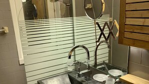 Un bany modern amb un lavabo negre, un aixeta d'acer inoxidable i un mirall. Hi ha una prestatgeria de fusta sobre el lavabo i una separació de vidre amb línies horitzontals.