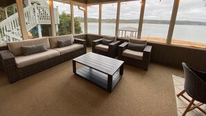 A cozy indoor seating area with a sofa, two armchairs, and a coffee table, overlooking a lake with a wooden deck and stairs in the background.
