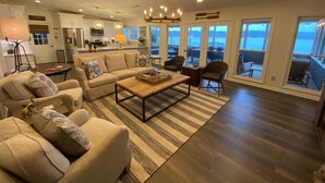 A spacious living room with a sofa, coffee table, and chairs, a kitchen in the background, and large windows offering a view of the outdoors.