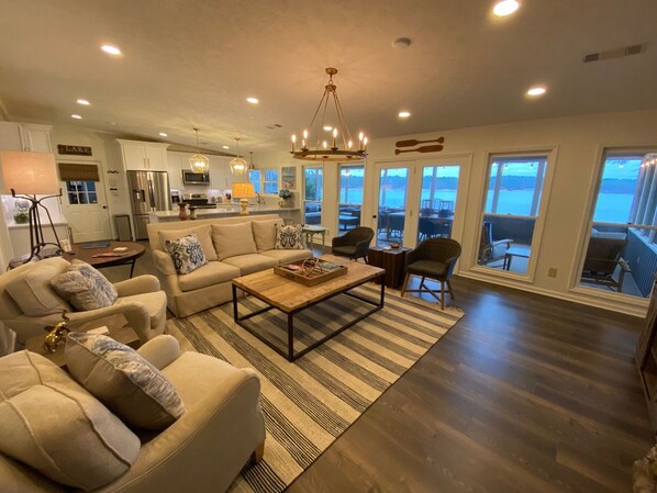 Un salón espacioso con sofá, mesa de centro y sillones, una cocina al fondo y ventanas amplias con vistas al exterior.