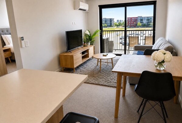A modern living room with a TV, sofa, dining table, and a view of the city through large windows. - Remarkables Park: Modern Apartment with Parking (Queenstown)