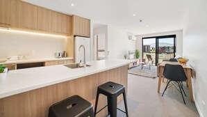 A modern kitchen with wooden cabinets, a white countertop, and a black stool. There is a dining area with a table and chairs, and a view of buildings through large windows. - Remarkables Park: Modern Apartment with Parking (Queenstown)