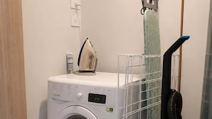 A laundry area with a front-loading washing machine, a drying rack, and a vacuum cleaner. - Remarkables Park: Modern Apartment with Parking (Queenstown)