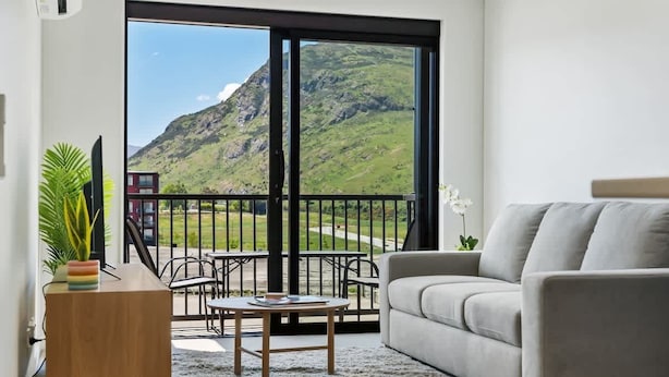 A living room with a grey sofa, a wooden side table, and a coffee table. A large window offers a view of a mountain and a red building. - Remarkables Park: Modern Apartment with Parking (Queenstown)