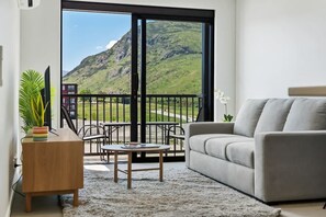 A living room with a grey sofa, a wooden side table, and a coffee table. A large window offers a view of a mountain and a red building. - Remarkables Park: Modern Apartment with Parking (Queenstown)