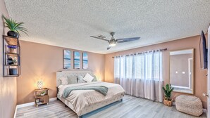 A bedroom with a bed, a ceiling fan, a nightstand, a window with curtains, and a wall-mounted shelf with plants.