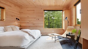 Een moderne slaapkamer met een bed, een leestoel, een bijzettafeltje en een groot raam met uitzicht op bomen.