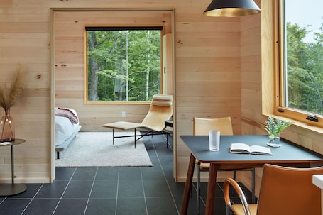 A cozy cabin interior with a wooden interior, a dining area with a table and chairs, a sofa, and a large window offering a forest view.