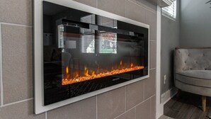 Een moderne woonkamer met een flatscreen televisie aan een betegelde muur, een grijze bank en een open haard met vlammen.