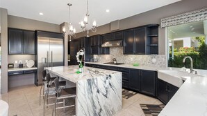 A modern kitchen with dark cabinets, a marble island, stainless steel appliances, and a large window overlooking greenery. - Villa Del Sol - 3 Suites w/ Pool, Spa, Firepit (La Quinta)
