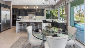 A modern kitchen with dark cabinets, stainless steel appliances, and a glass island with seating. - Villa Del Sol - 3 Suites w/ Pool, Spa, Firepit (La Quinta)