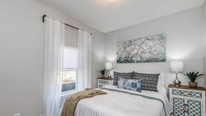 Un dormitorio con una cama grande, cortinas blancas, un cuadro colgado en la pared, lámparas de noche y un aparador decorativo.