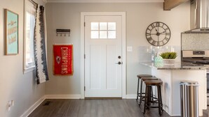A modern kitchen with a white door, a clock, bar stools, and a stainless steel trash can. - West Plaza House in the heart of KC (Kansas City)