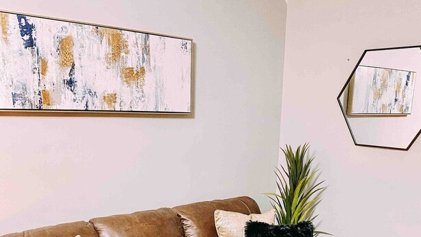 Ein braunes Ledersofa mit zwei schwarzen, flauschigen Zierkissen, ein kleiner Beistelltisch mit einem Buch und ein großes abstraktes Gemälde an der Wand.