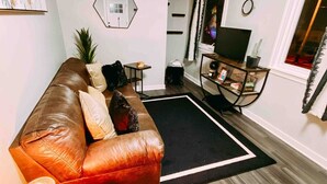 A living room with a brown leather sofa, a small wooden side table, a black rug, and a television on a stand. - West Plaza House in the heart of KC (Kansas City)