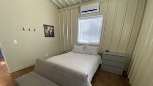 Un dormitorio con cama, mesitas de noche, aire acondicionado, ventana con persianas, un cuadro en la pared y una puerta.
