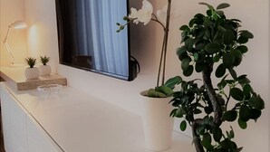 Ein modernes Wohnzimmer mit einem flachbildfernseher an der Wand, einem kleinen Tisch mit Lampe und dekorativen Pflanzen, sowie einem Blumentopf auf einem weißen Konsolentisch.