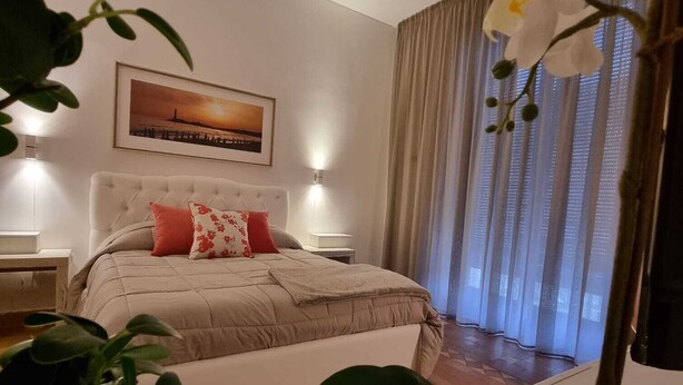 Ein Schlafzimmer mit einem Bett, einem Nachttisch, einer Wandlampe, einem Fenster mit Vorhängen und einem gerahmten Bild an der Wand.