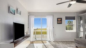 Een woonkamer met een flatscreentelevisie, een plafondventilator, een groot raam met gordijnen en uitzicht op een groen landschap.