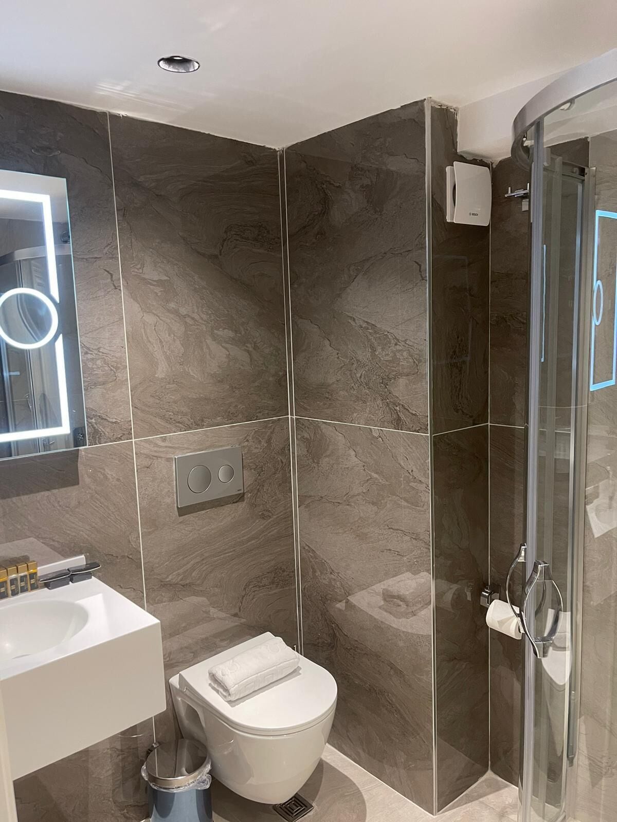 A modern bathroom with a white toilet, a glass shower enclosure, and a large mirror with a circular light.