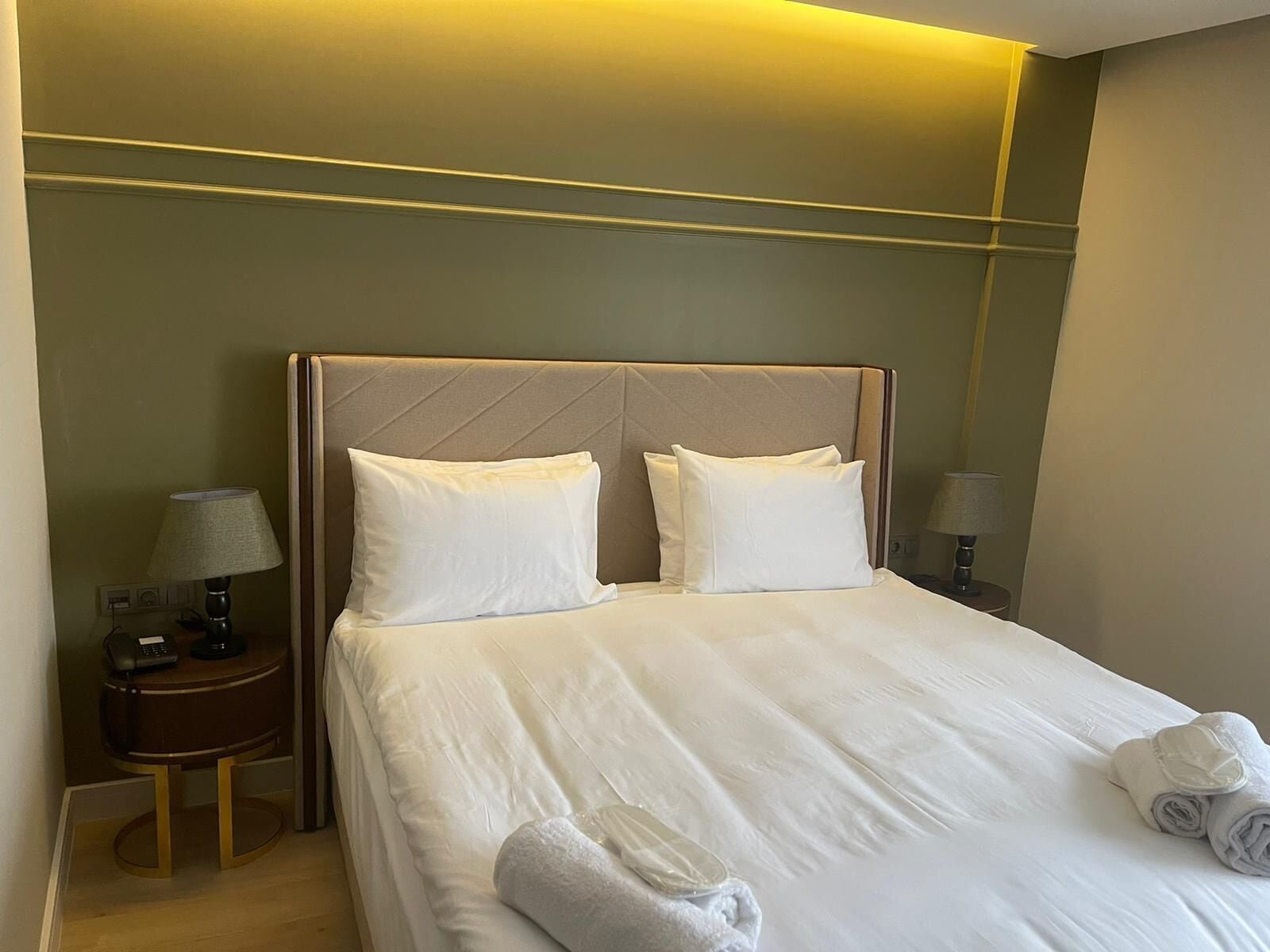 Habitación de hotel con una cama grande, dos almohadas, una mesita de noche con una lámpara y una toalla enrollada sobre la cama.