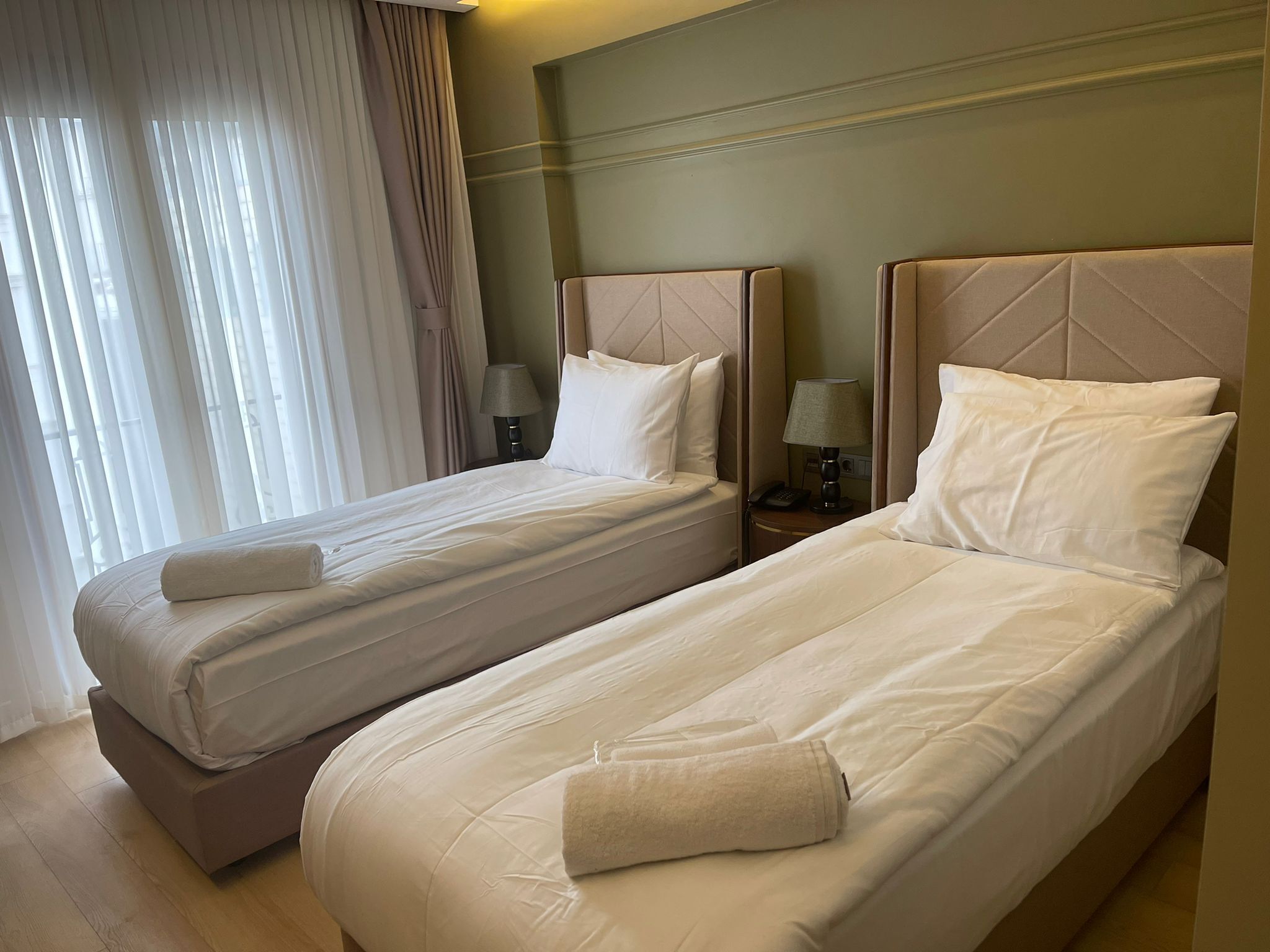 Une chambre d’hôtel avec deux lits simples, chacun doté d’une tête de lit, de tables de chevet avec des lampes et d’une fenêtre avec des rideaux légers.