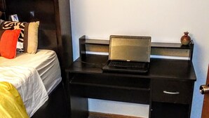 Quarto com cama, escrivaninha, laptop, pequeno vaso e porta.